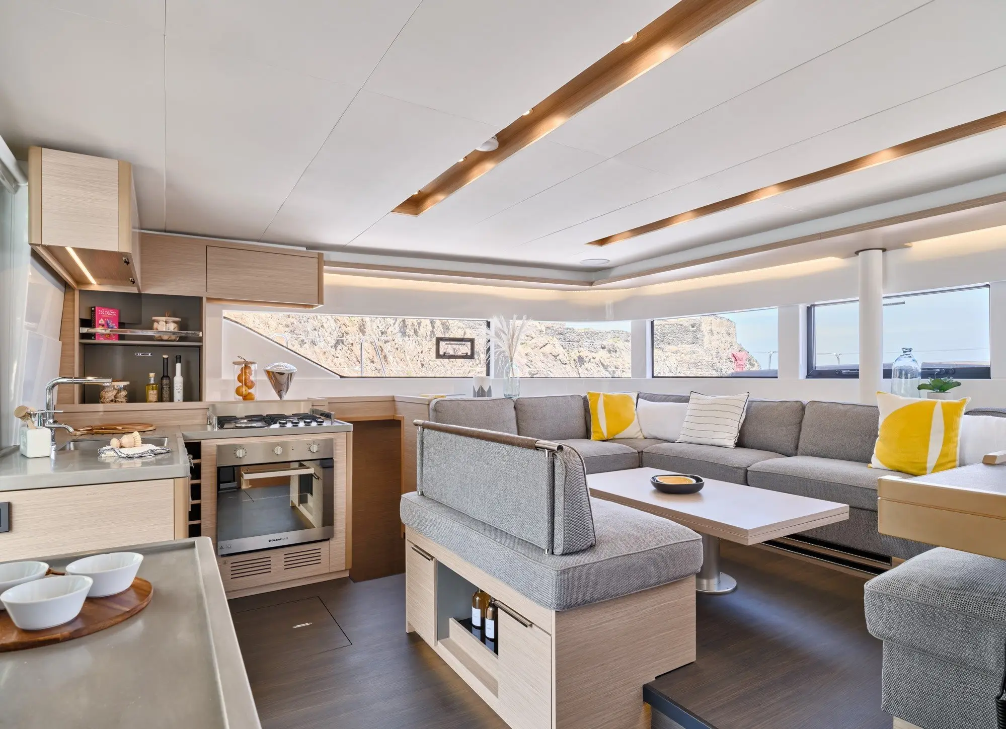 Modern Lagoon 51 yacht interior with large windows, light wood cabinetry, a kitchen, and a gray L-shaped sofa with yellow and white pillows. Natural light fills the spacious, stylish living area with a scenic view outside.