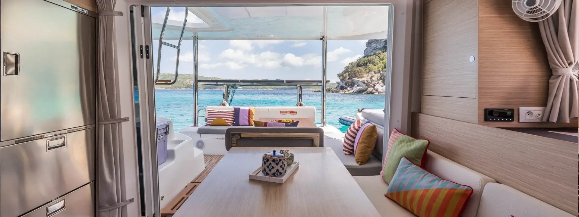 View from inside a modern Lagoon 38 boat cabin with a dining table and colorful cushions, looking out to turquoise water, cliffs, and a blue sky through open doors to the deck.