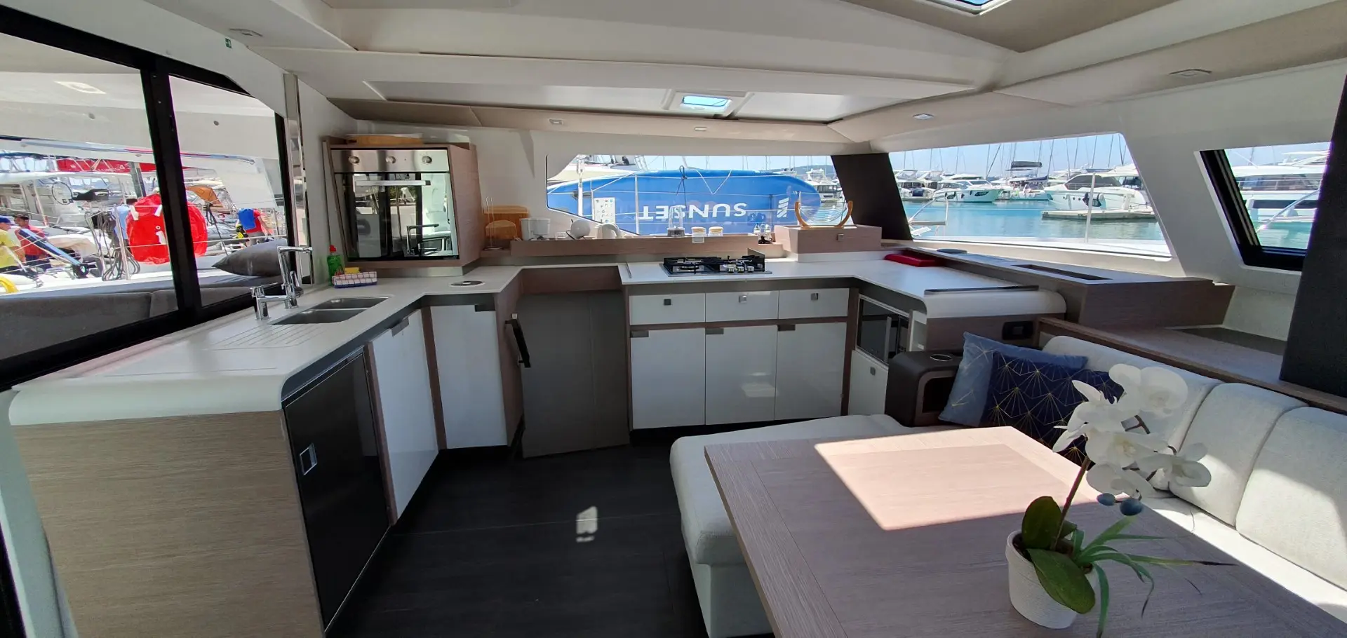 Modern yacht interior with a bright kitchen, dining area, white cabinets, and large windows overlooking a marina with boats and a blue SUNSELF sign outside. A potted orchid is on the table aboard the Fountaine Pajot Elba 45.