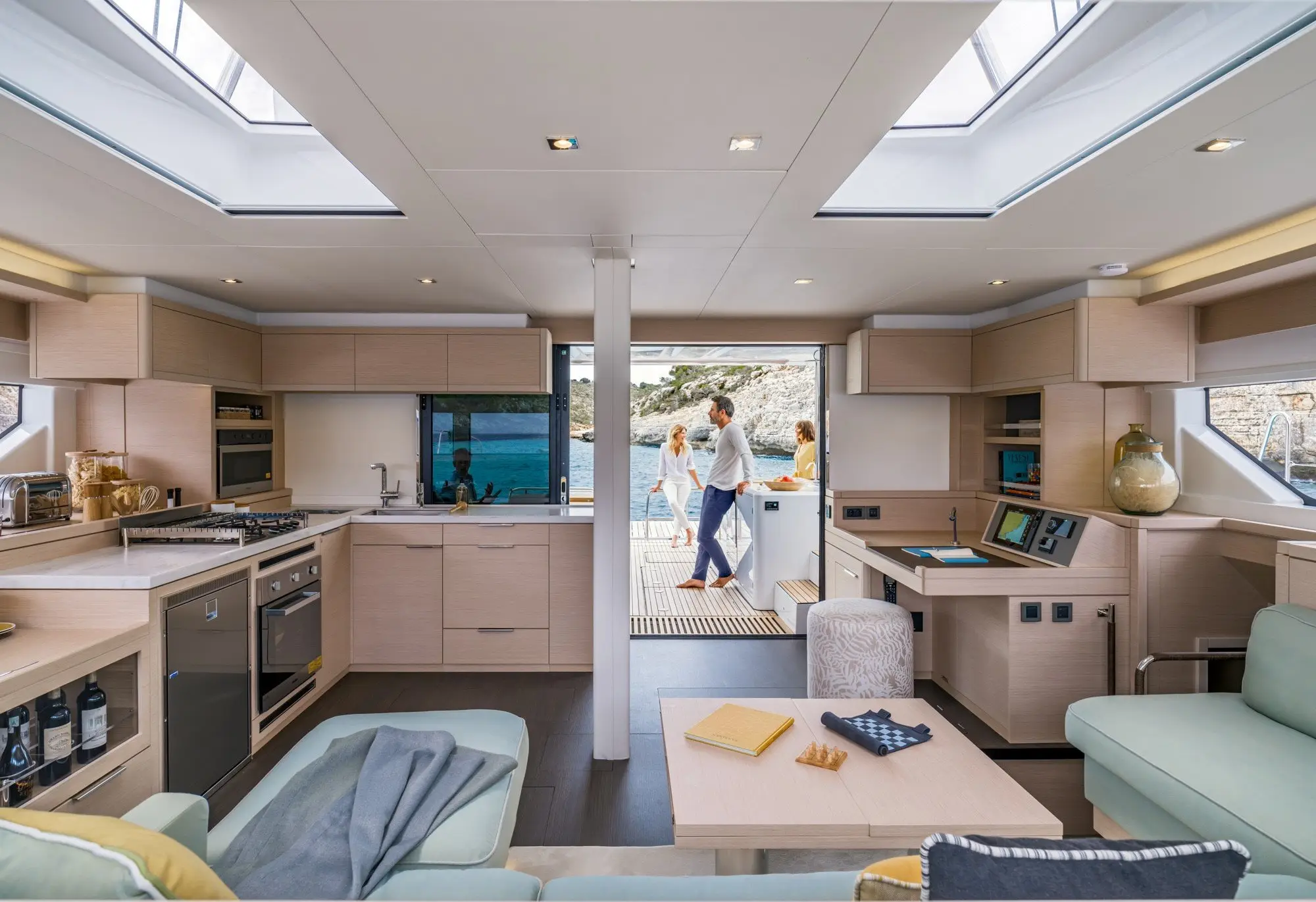 Modern Lagoon 55 yacht interior with light wood finishes, kitchen and lounge area, large windows, and a view of people standing outside on the deck by the sea. Natural light fills the spacious, stylish cabin.