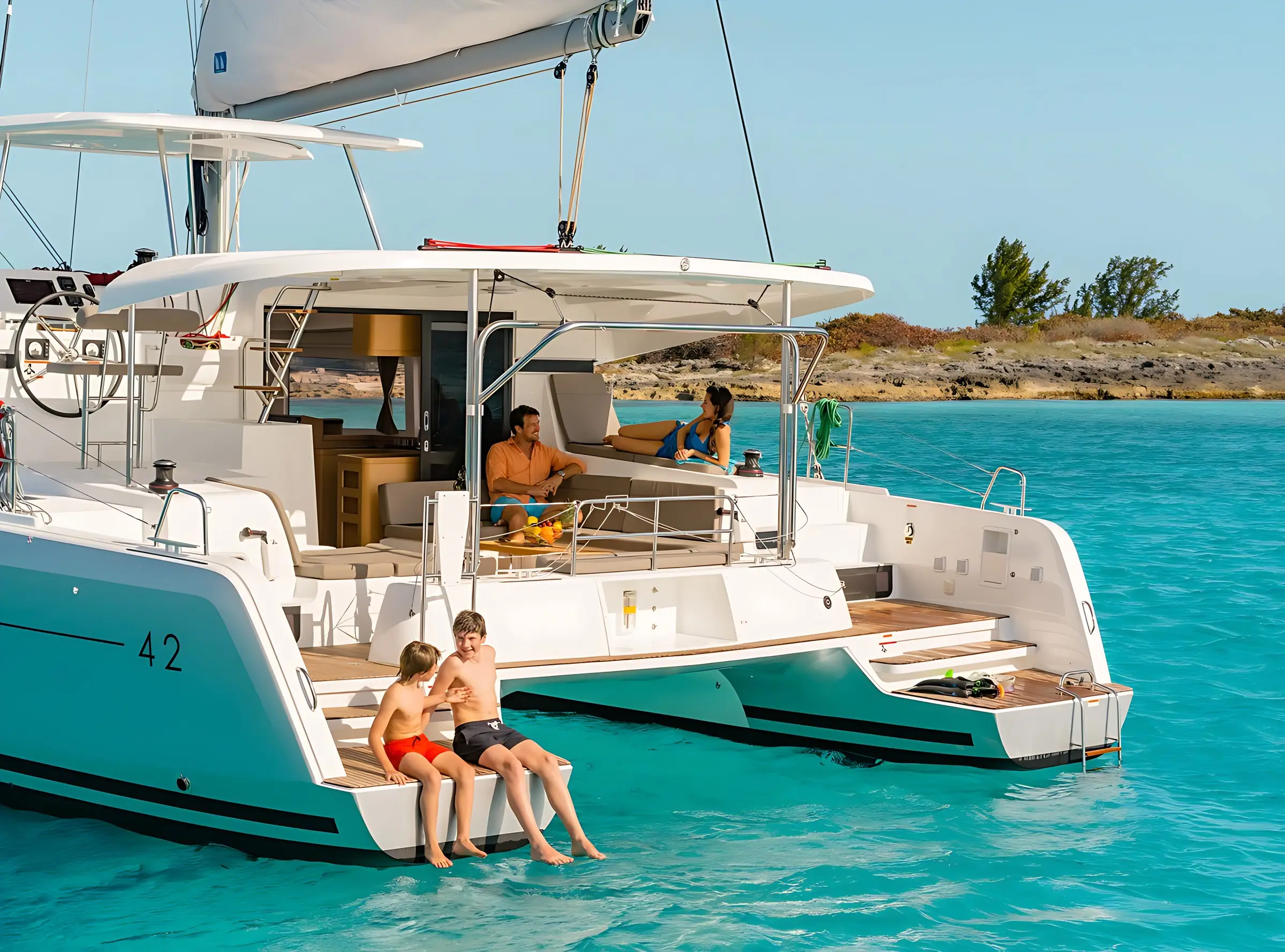 Four people relax on a white catamaran yacht in clear blue water; two sit at the back with their feet in the water, while two others lounge and talk under the shaded seating area. Rocky shoreline and trees are visible in the background.