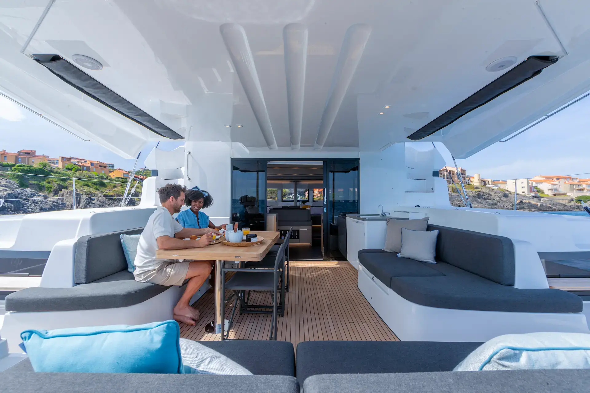 Two people sit at a table on the deck of a Lagoon 51 yacht, enjoying drinks and food. The yacht features sleek gray seating, a wooden floor, and ocean views with buildings visible on the coastline in the background.