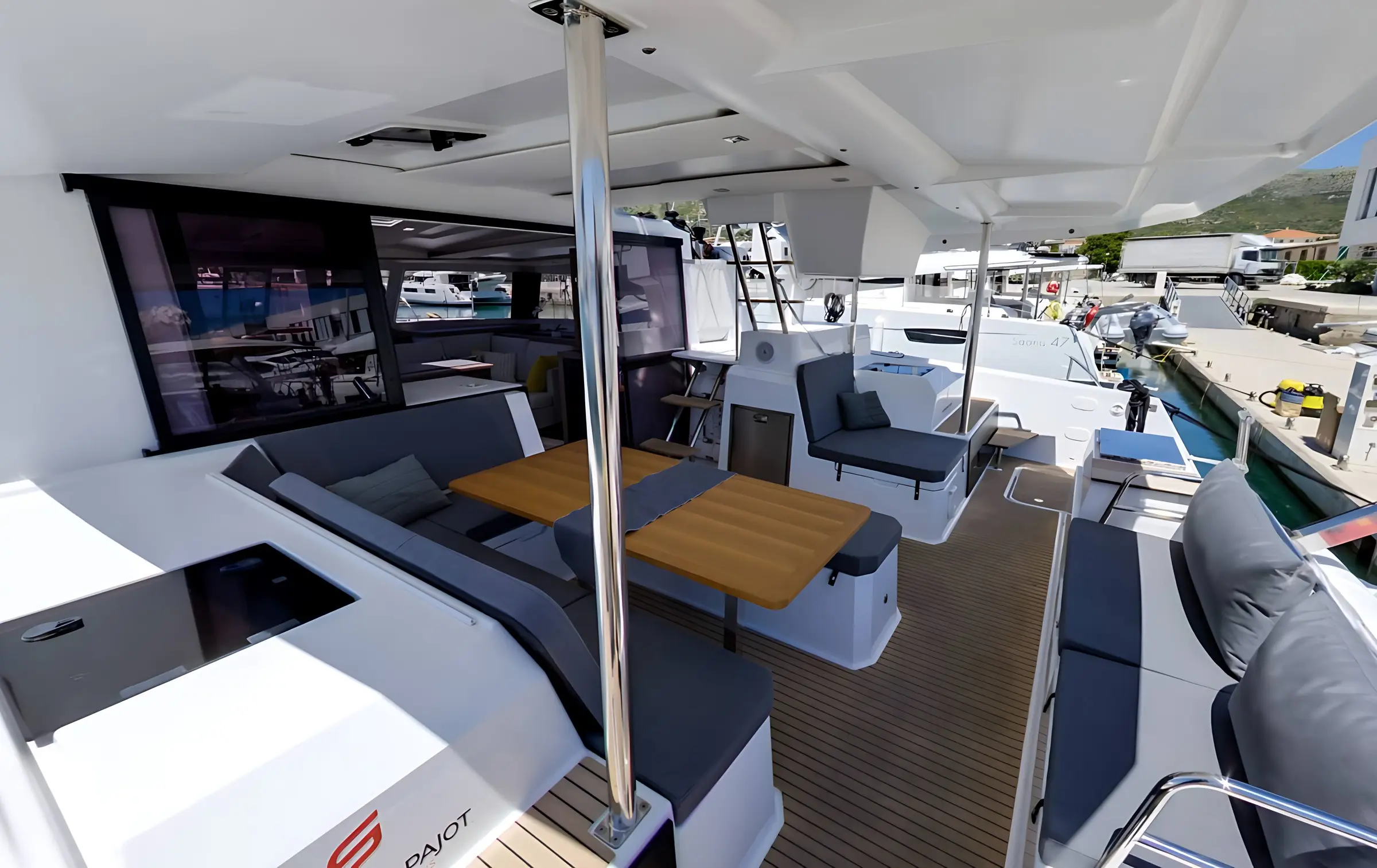 The image shows the outdoor seating area of a modern Fountaine Pajot Astrea 42 yacht with grey cushioned benches, a wooden table, and a shaded canopy. The yacht is docked at a marina, with other boats and a building visible in the background.
