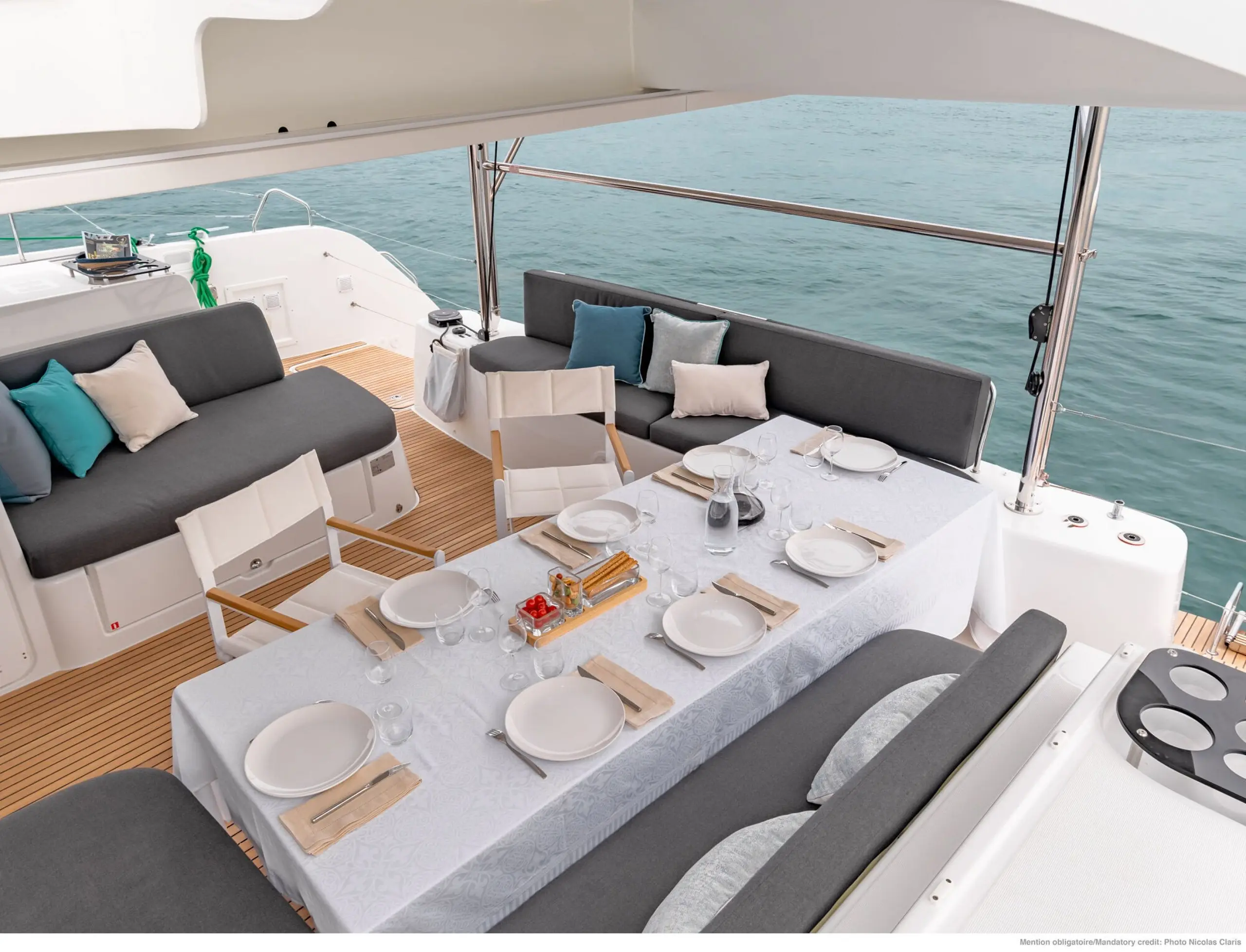 A luxurious Lagoon 46 yacht deck features a dining table set for eight with white tablecloth, plates, glasses, and napkins. Gray cushioned seats and throw pillows surround the table, overlooking calm blue water.