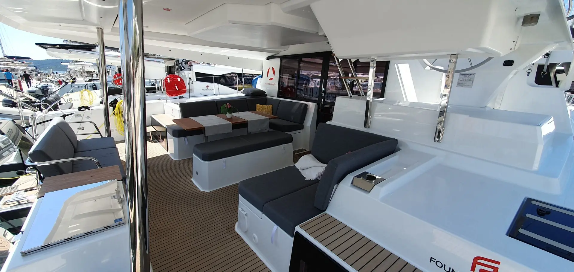 The image shows the deck of a modern Fountaine Pajot Elba 45 catamaran yacht, featuring cushioned seating, a table with plants and drinks, and wooden flooring. Other boats and a marina can be seen in the background.