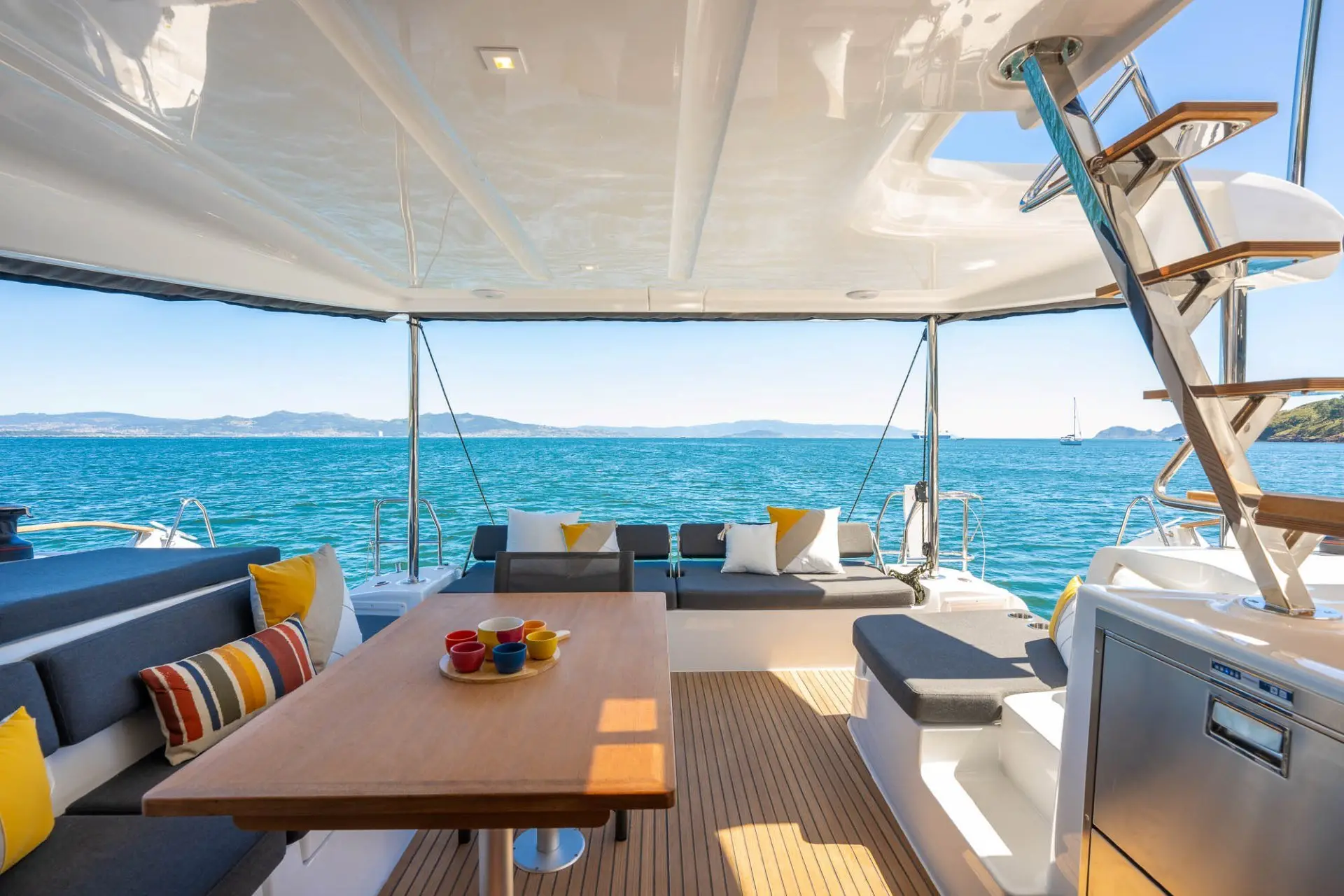 Spacious Lagoon 43 yacht deck with a wooden table, colorful cups, cushioned seating, and decorative pillows. The deck overlooks calm blue water and distant hills under a clear sky. Stainless steel stairs lead to an upper level.