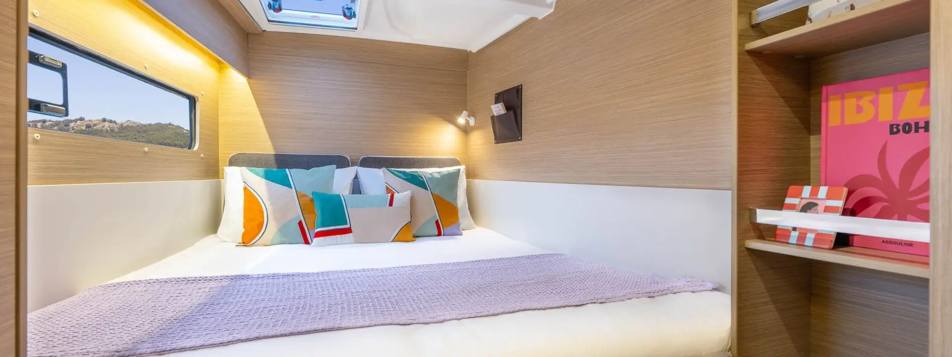A cozy cabin bedroom on a Lagoon 38 with a double bed, colorful geometric pillows, a lavender throw, overhead lights, a scenic window view, and a shelf holding books—including one with an IBIZA cover.