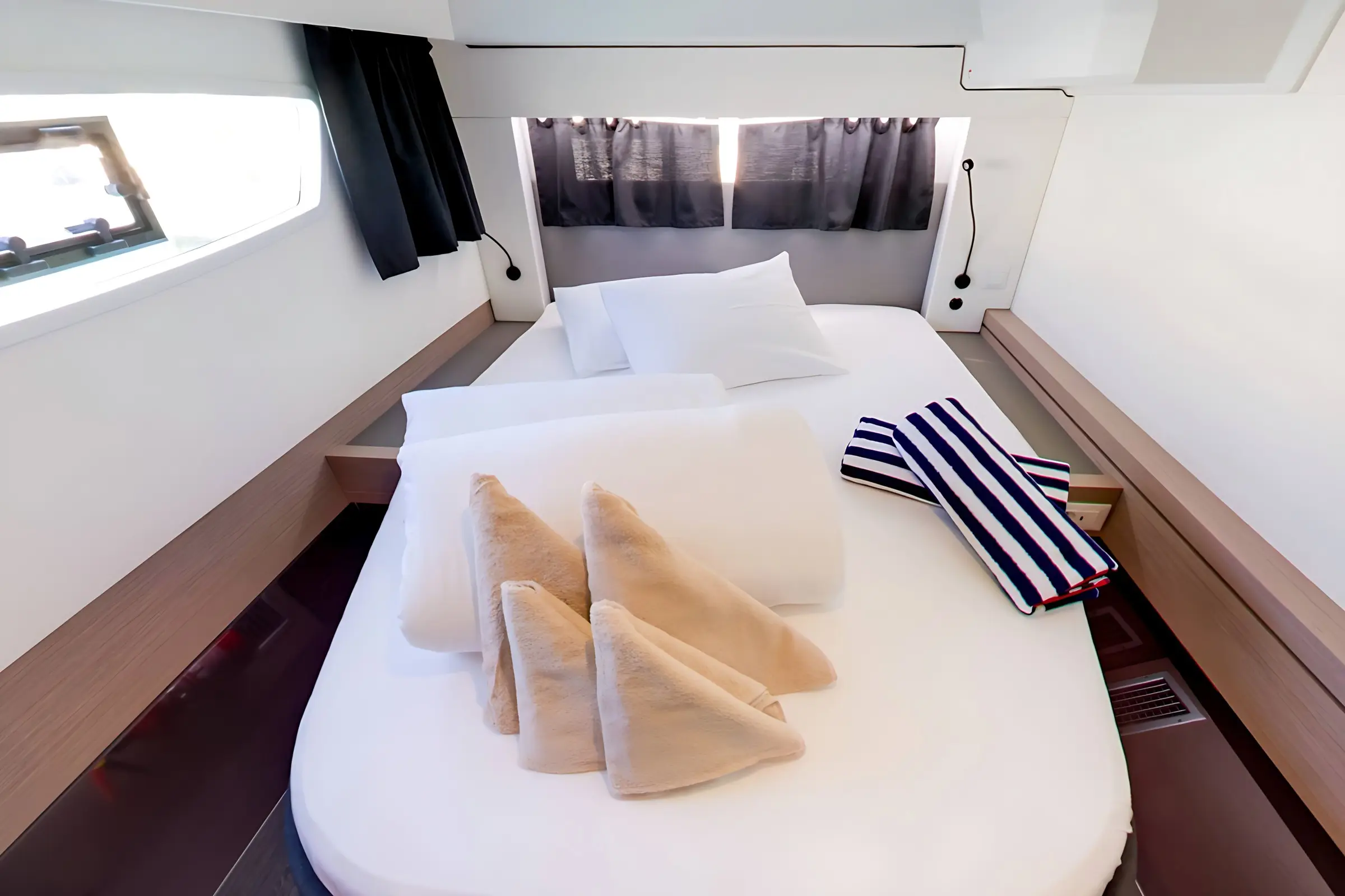 A neatly made bed in a compact room aboard the Fountaine Pajot Astrea 42 features white bedding, three beige folded towels, and two striped beach towels. The space includes wood accents, large windows with black curtains, and a pillow propped against the headboard.