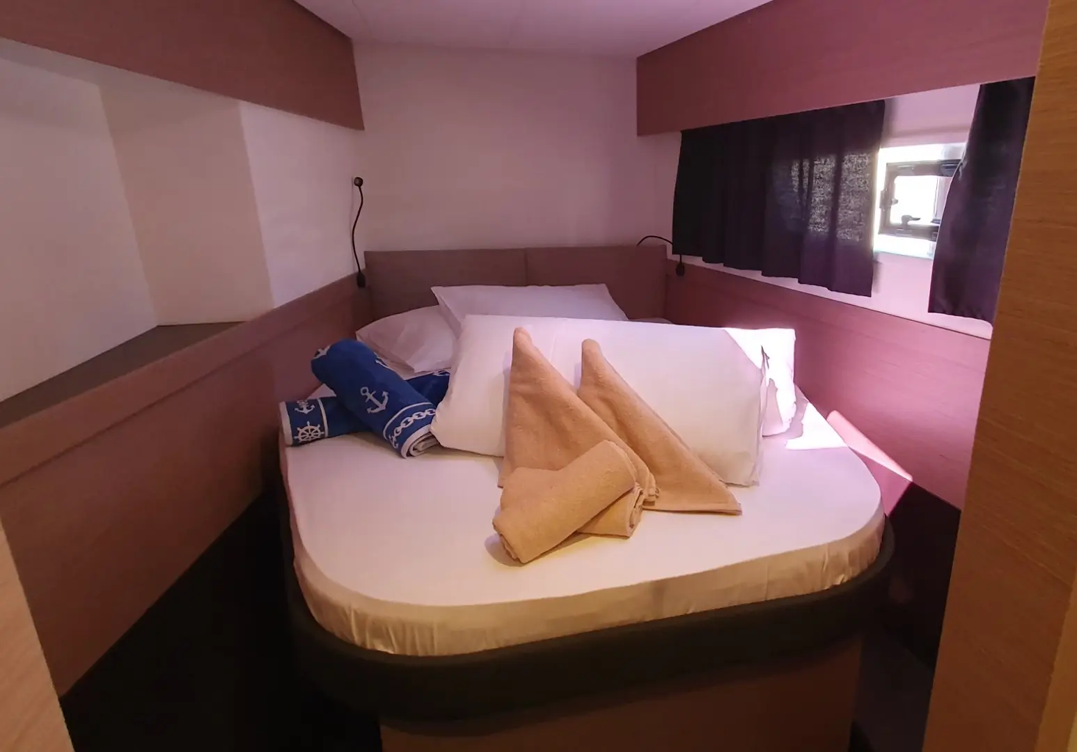 A cozy bedroom aboard the Fountaine Pajot Elba 45 features a made-up double bed, two folded towels shaped like sailboats, white pillows, and rolled blue towels with nautical designs. Soft light streams through a small window with a black curtain.