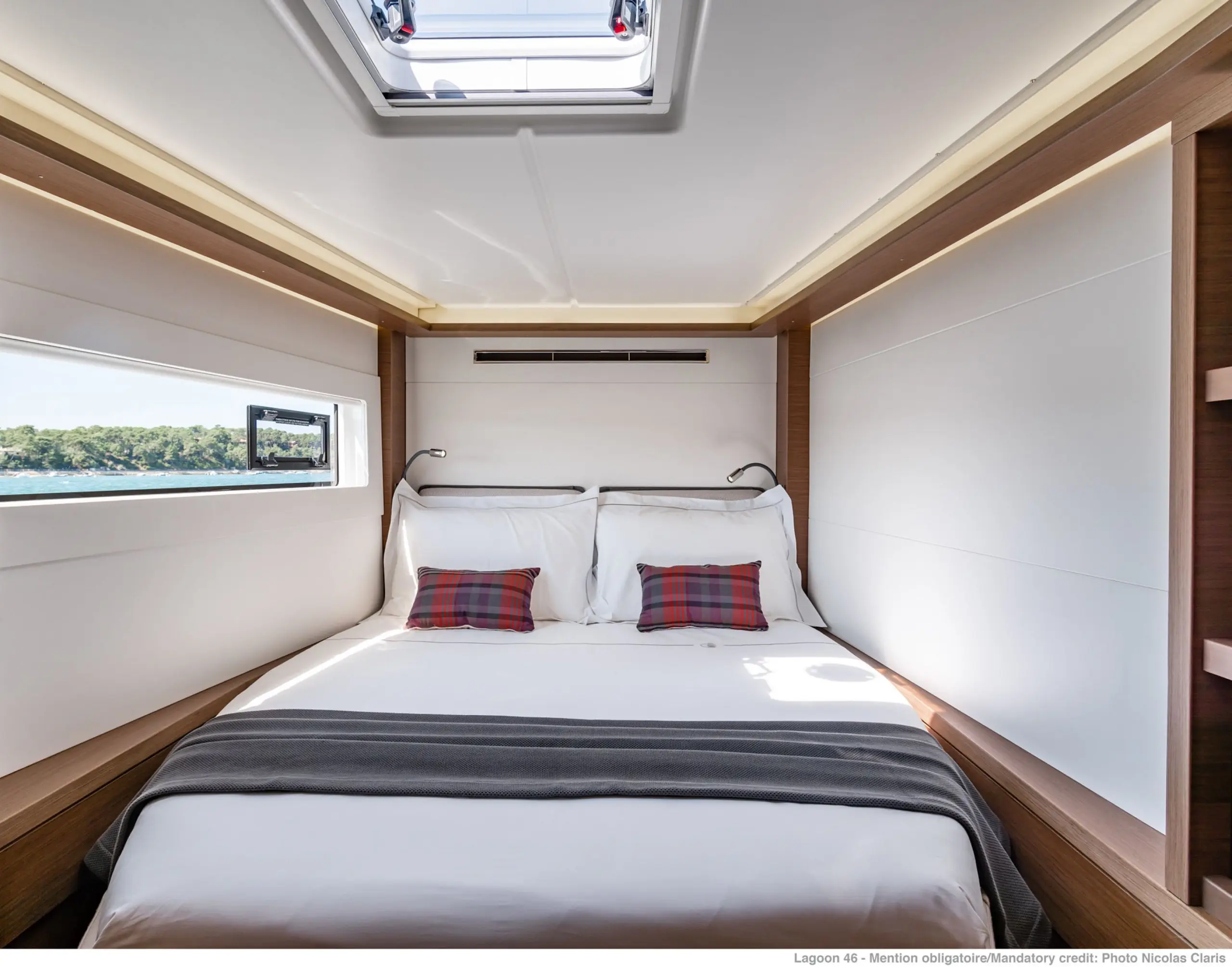 A modern Lagoon 46 boat cabin with a neatly made double bed, two plaid accent pillows, a large window on one side, a skylight above, and wood paneling along the walls. Natural light fills the cozy, minimalist space.