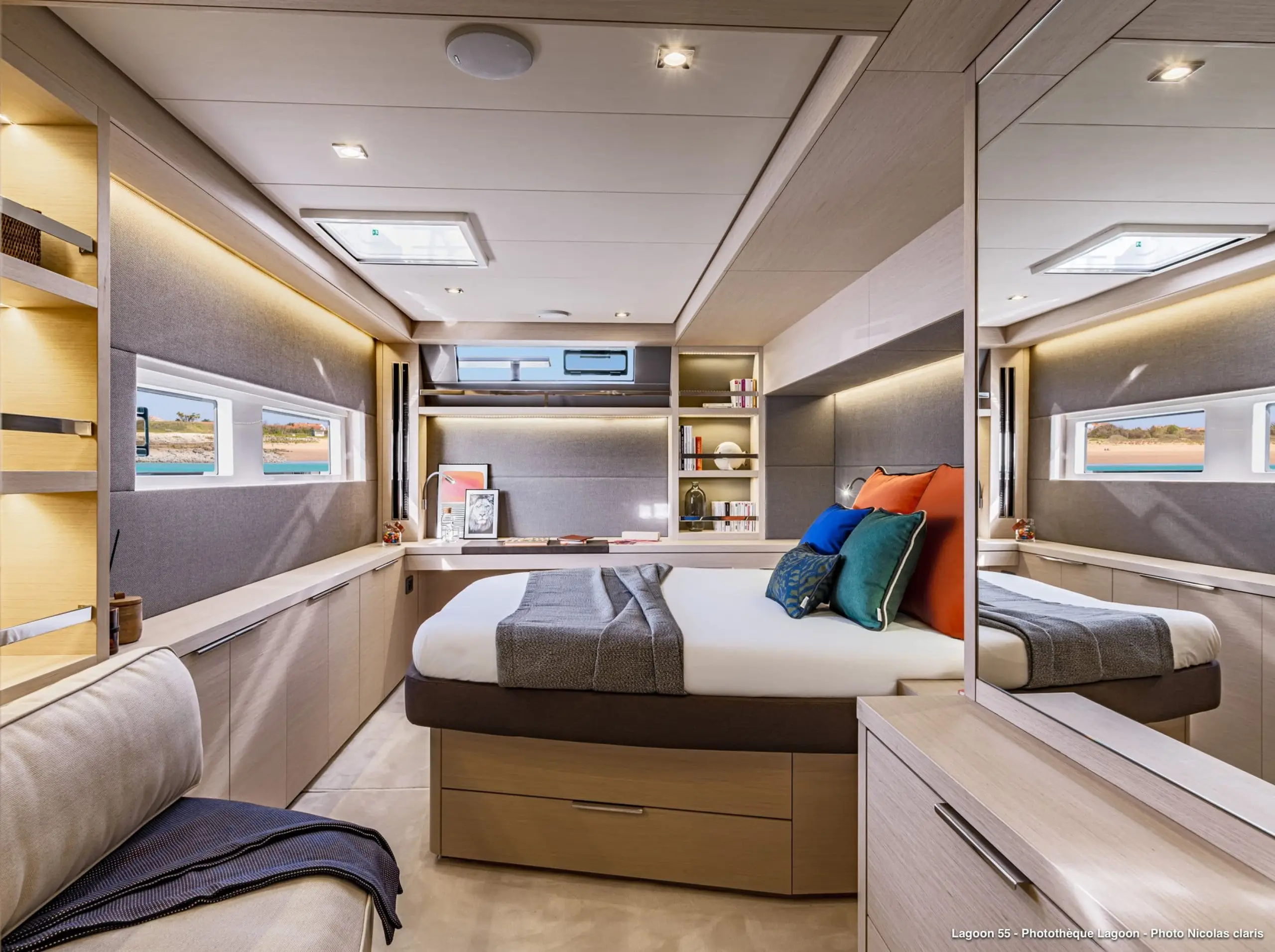 Modern bedroom with a large bed, colorful pillows, built-in shelves, desk, and windows letting in natural light—echoing the relaxed elegance of the Lagoon 55. Light wood furniture and soft lighting create a cozy, inviting atmosphere.