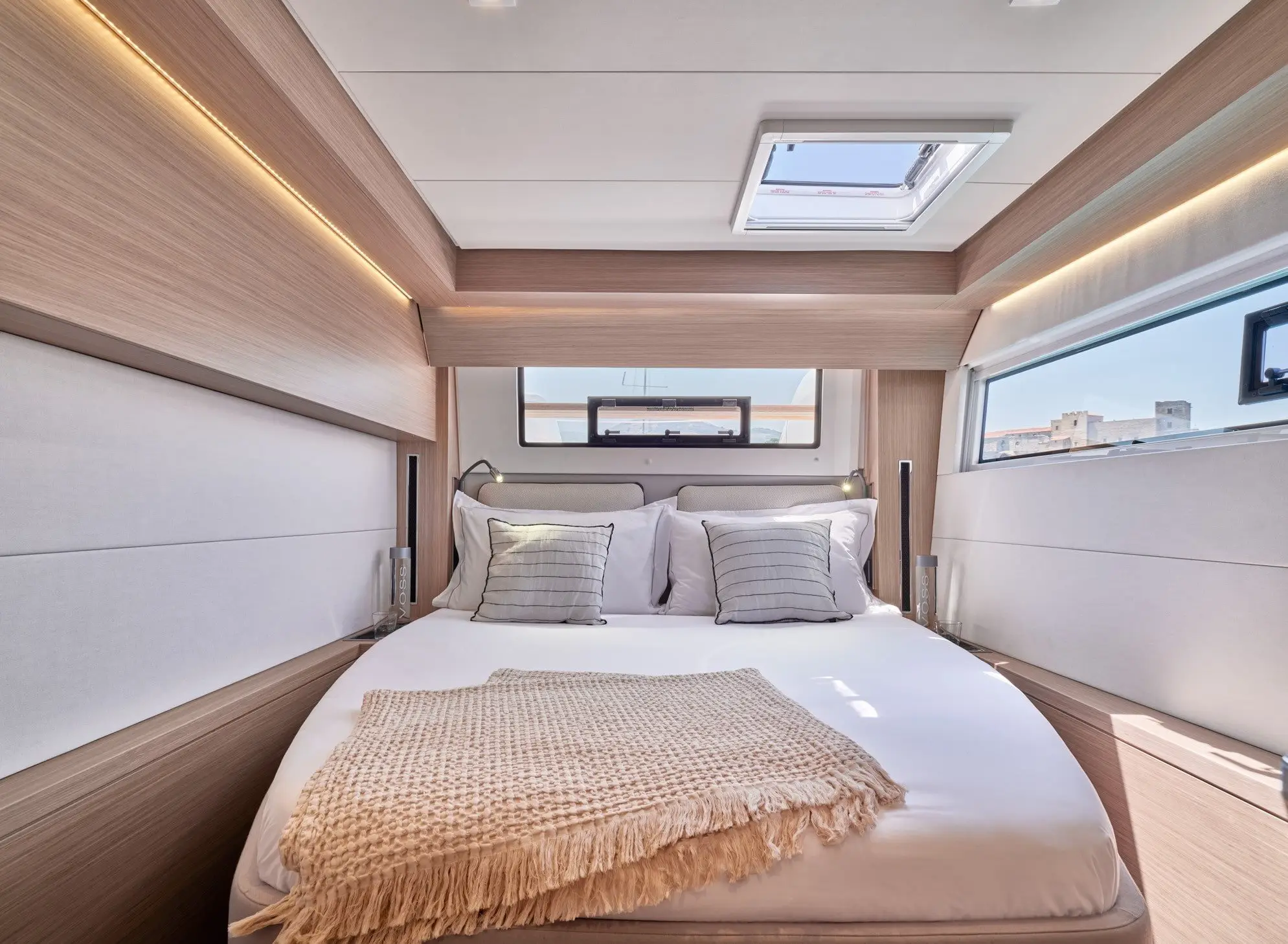 A cozy Lagoon 51 yacht cabin featuring a neatly made bed with white linens, four pillows, a beige throw blanket, wood paneling, large windows, and a skylight allowing natural light to fill the room.