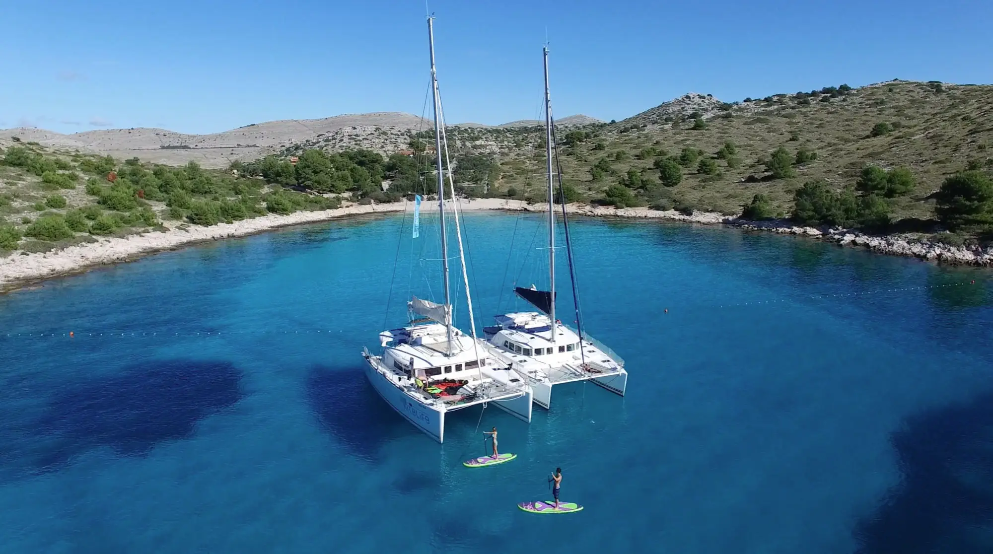 Two catamarans anchored in a clear blue bay near a rocky, green shoreline. Two people paddleboard on the calm water between the boats and the shore under a sunny sky.