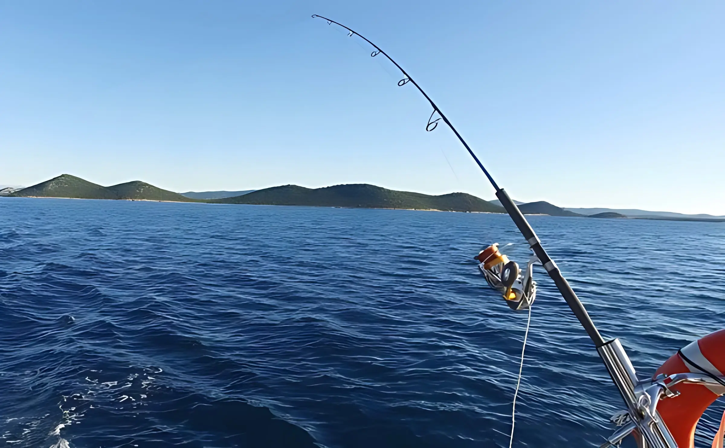 A fishing rod is mounted on a boat over deep blue water, with distant green hills and a clear sky in the background—an ideal scene for catamaran activities.