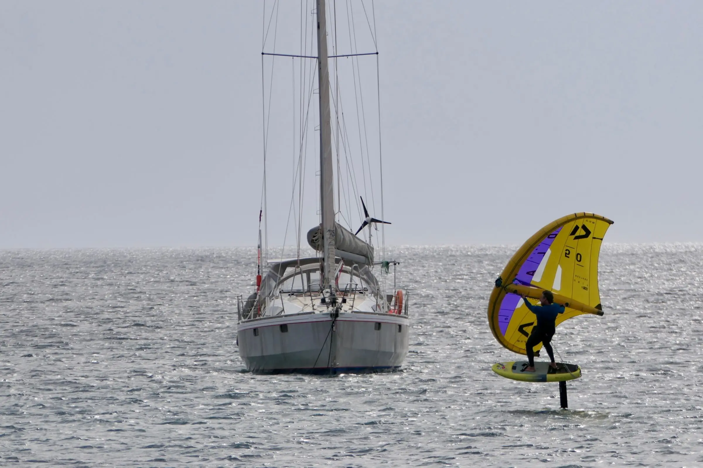 Wing foiler with a yellow wing riding past an anchored sailing yacht
