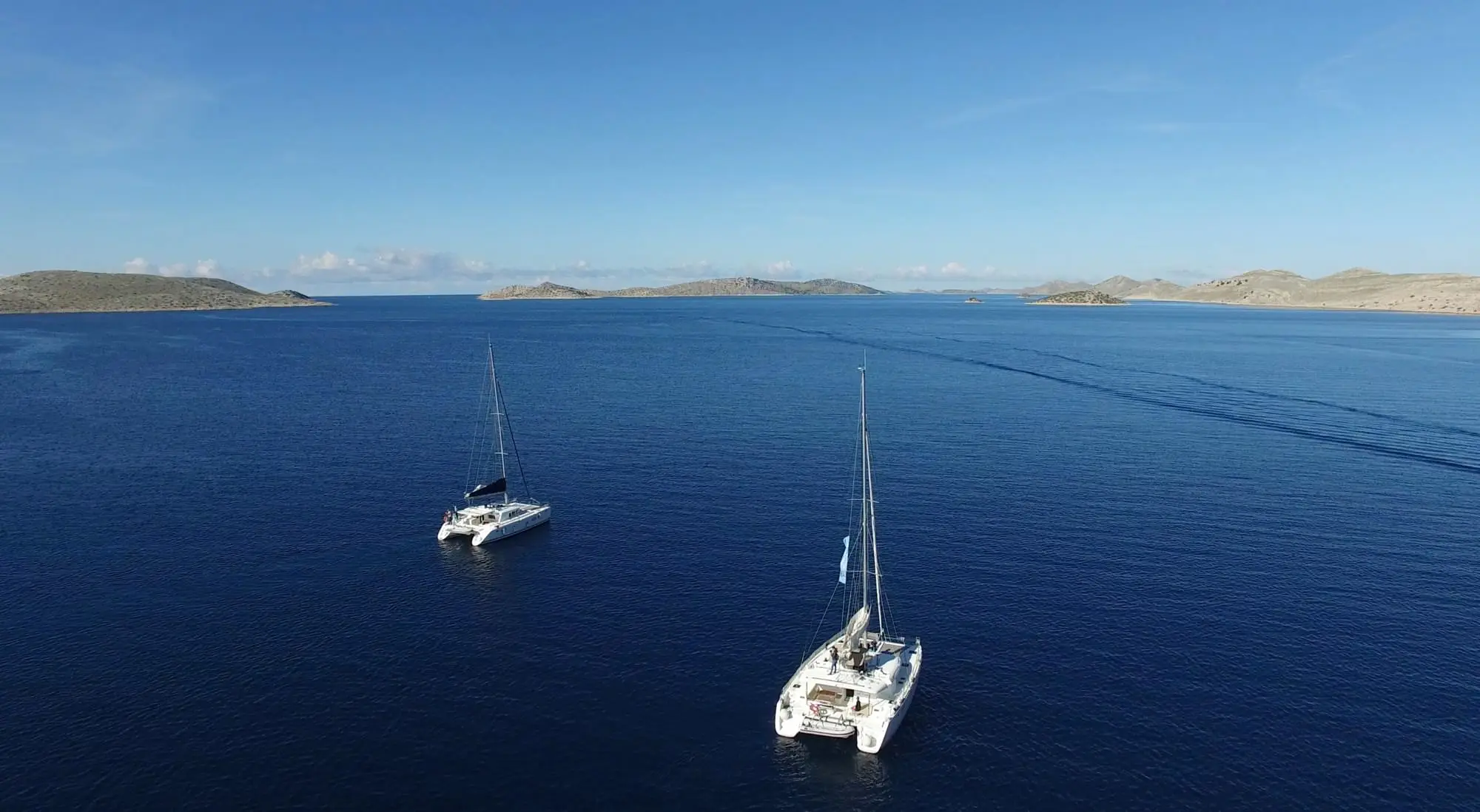 Two sailboats on calm, deep blue water under a clear sky, small islands in the distance.